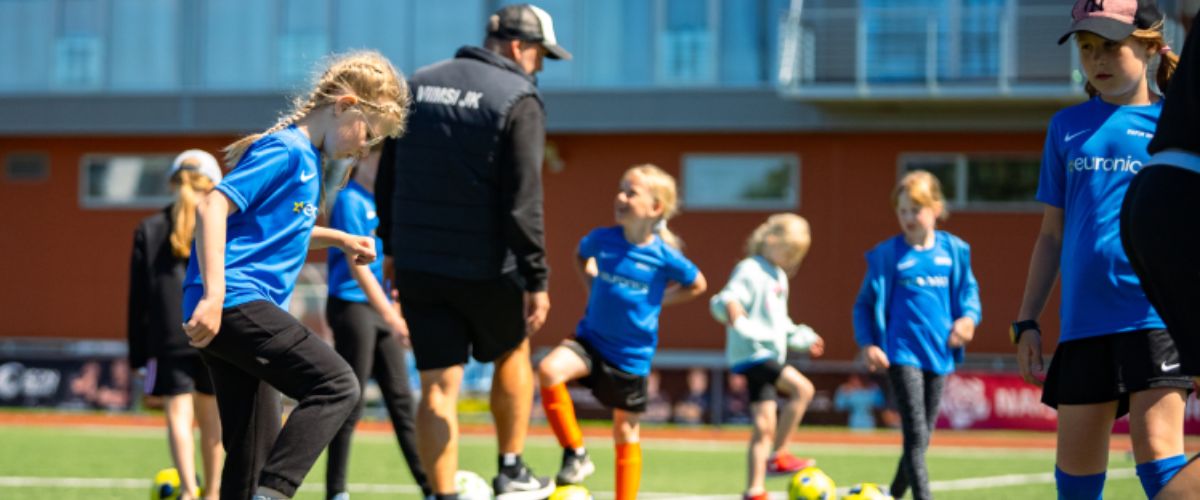 Tüdrukud Euronicsi jalgpallilaagris harjutamas palliga mängu, taustal treener juhendamas ja teised lapsed aktiivselt kaasa löömas. Lapsed kannavad siniseid Euronicsi treeningsärke ning tegevus toimub kunstmuruväljakul päikeselisel päeval.