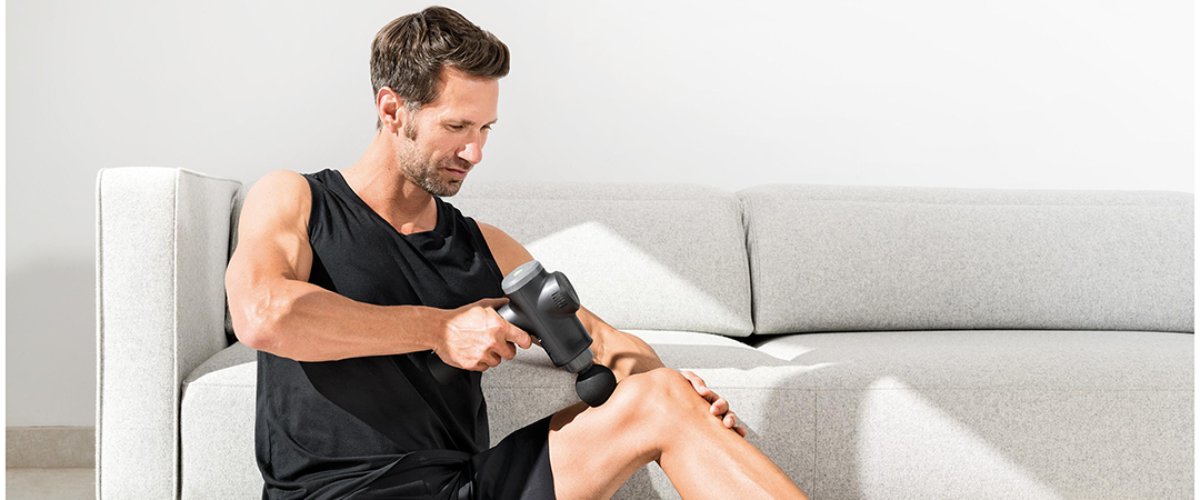Мужчина использует массажёр Beurer для расслабления мышц ног, сидя рядом с диваном