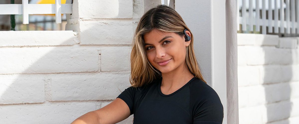 Naeratav naine valge seina taustal kõrvas juhtmevabad JLab Sport Air spordikõrvaklapid