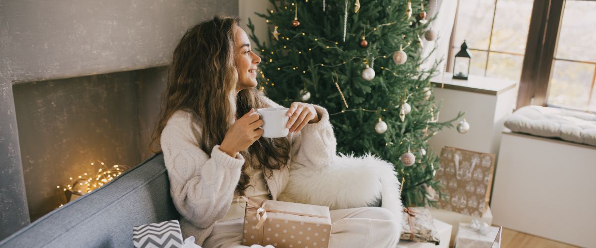 Женщина сидит на диване рядом с елкой и держит чашечку, рядом с ней — упакованный рождественский подарок для женщины