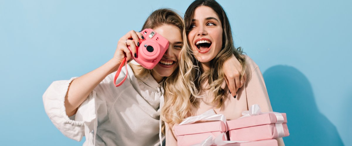 Kaks rõõmsat naist teevad kiirpildikaameraga selfiet ja hoiavad roosasid kingikarpe