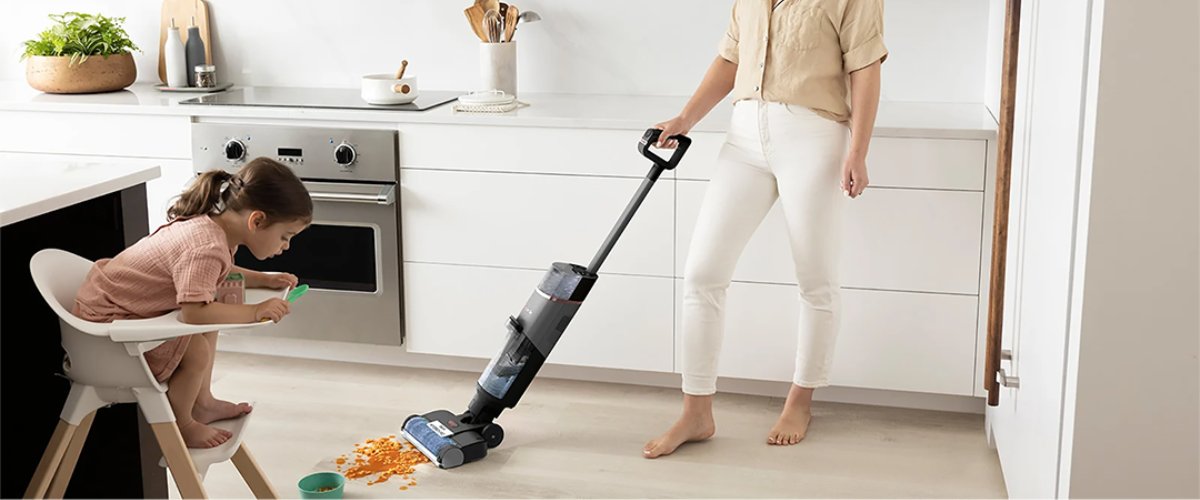 Naine koristab köögipõrandat juhtmevaba peseva tolmuimejaga, samal ajal kui väike laps istub söögitoolis ja ajab toitu maha