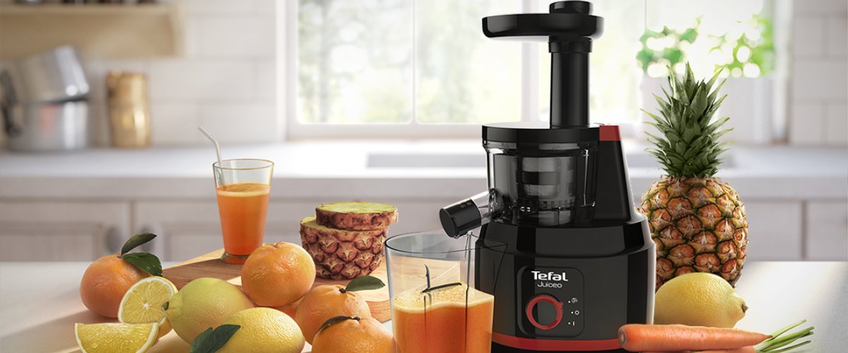 Шнековая соковыжималка Tefal Juiceo на кухонной столешнице, рядом свежий сок, цитрусовые, ананас и морковь